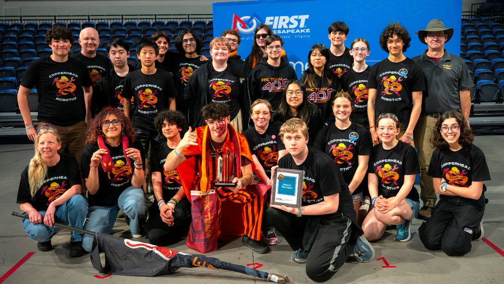 A large group of students and mentors from "Copperhead Robotics" pose with awards in front of a FIRST Chesapeake banner. Many wear black team t-shirts.