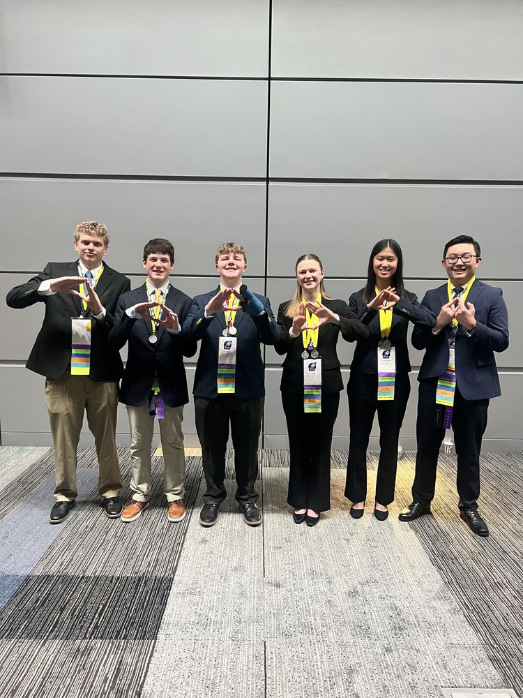  Gemini said Six students from the Blacksburg DECA chapter stand in a line against a gray paneled wall, celebrating their success at the State Leadership Conference. All six are dressed in professional business attire, including blazers and slacks, and wear yellow lanyards with competition credentials and colorful merit ribbons. Each student is using their hands to form a diamond shape, a signature DECA gesture, in front of their chest. The group includes four young men and two young women, all smiling as they pose on a multi-toned gray carpeted floor.