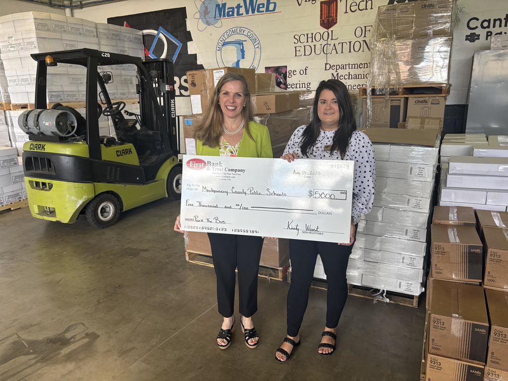 A medium shot shows two women in a warehouse holding a large check. The woman on the left has long, light brown hair and is wearing a green jacket over a white top and black pants. The woman on the right has long, dark brown hair and is wearing a black-and-white polka dot shirt and black pants. They are smiling at the camera. The check is from Union Bank & Trust Company and is for $5,000 made out to Montgomery County Public Schools. In the background, there are pallets of boxes, a yellow forklift, and a banner that reads Virginia Tech School of Education, Department of Mechanical Engineering.