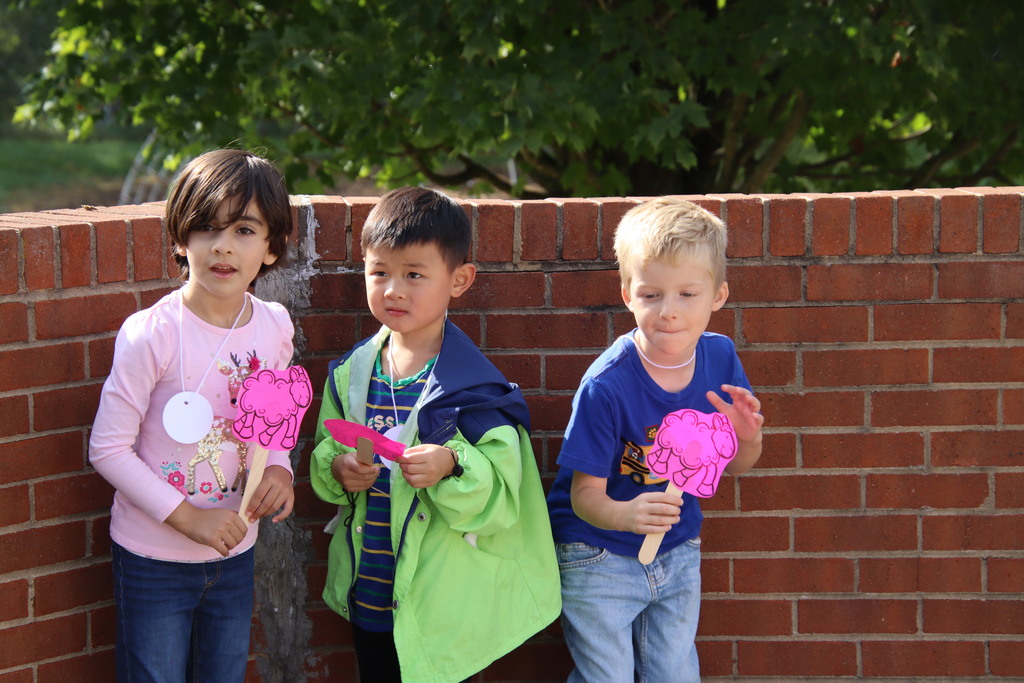 Second image: Three children stand in front of a brick wall, each holding pink paper cutouts of sheep on sticks. They are wearing casual clothes and look engaged in an outdoor activity.