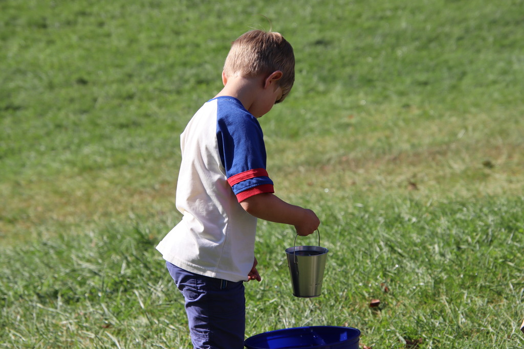 First image: A young boy in a white and blue T-shirt with red stripes on the sleeves is outdoors on grass, holding a small metal bucket near a larger blue container.