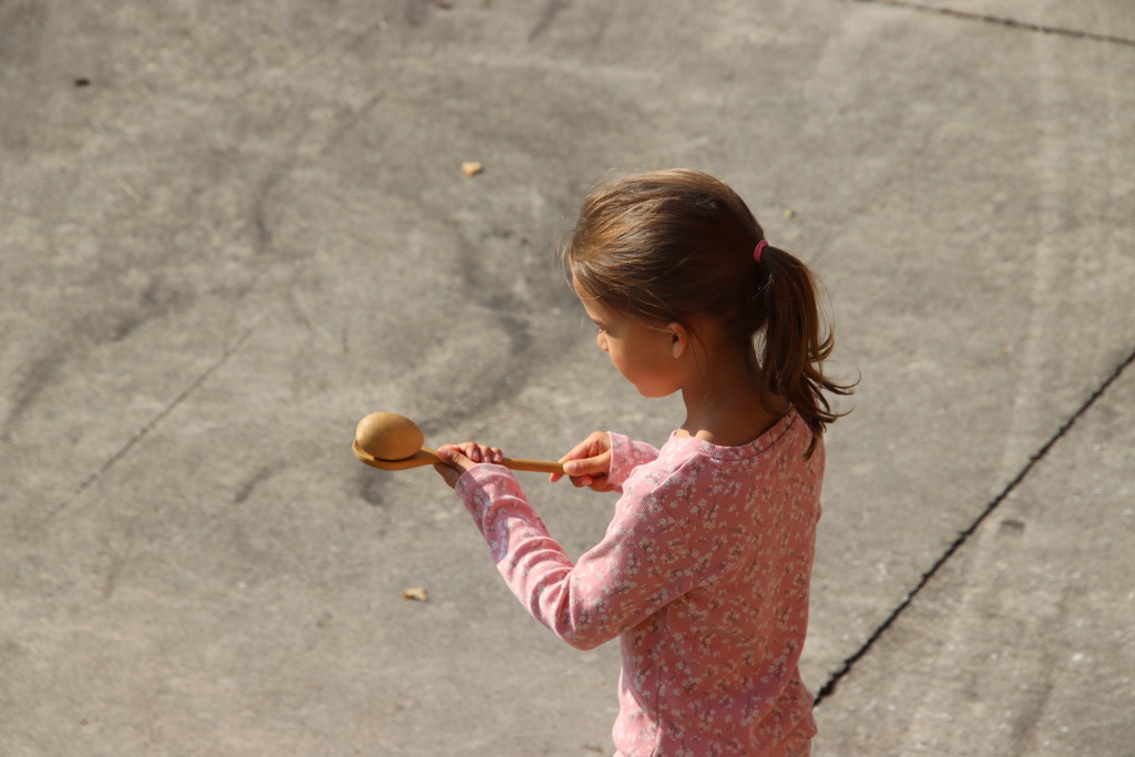 Third image: A young girl with her hair in a ponytail is outdoors holding a wooden spoon with a ball balanced on it, focusing intently as she participates in a game.
