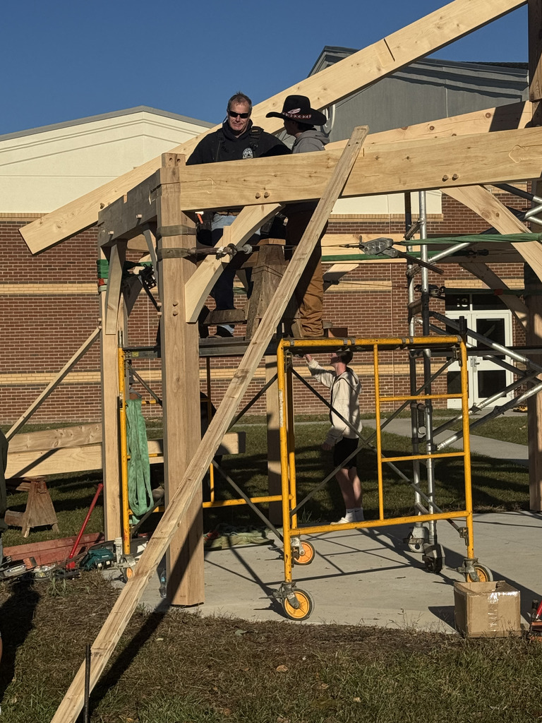 A close-up of two adults standing on scaffolding, fastening a wooden beam to a large vertical post on the timber frame structure. A youth stands below on a lower level of yellow scaffolding, assisting with the construction.