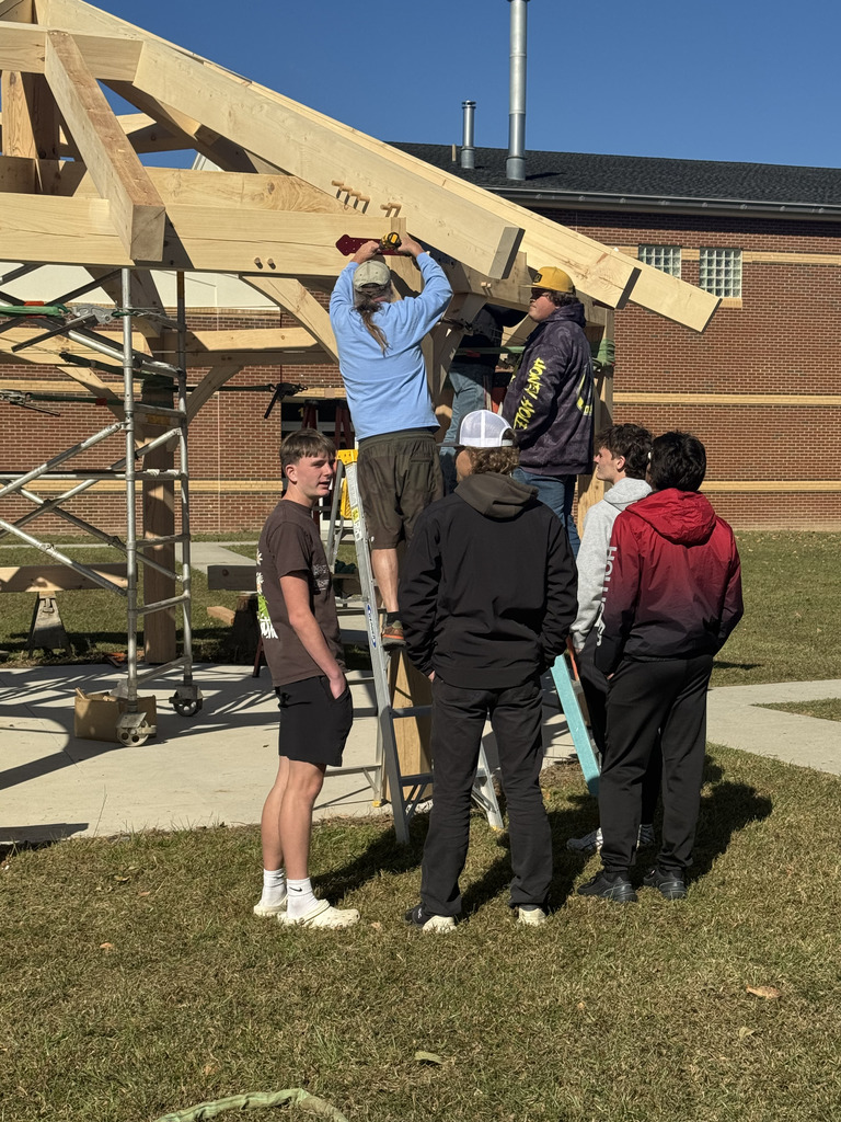 A group of young people, some standing on a scaffold and ladder, are working together to secure the wooden beams of a timber frame structure outdoors on a sunny day. A red brick building is in the background.