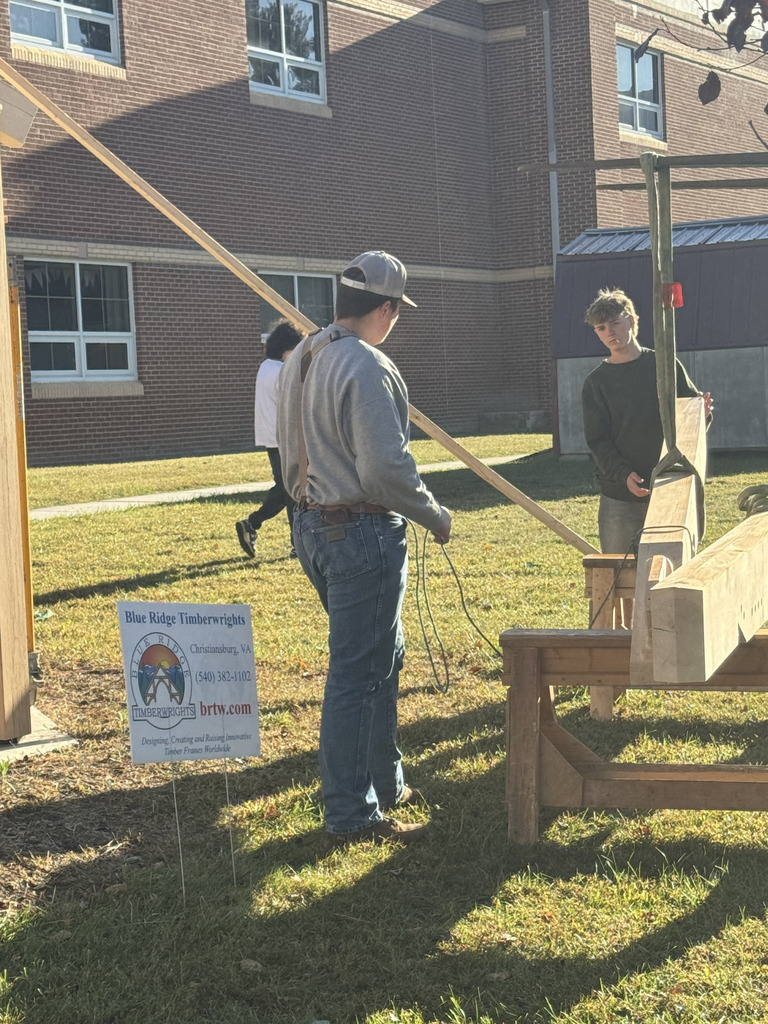A close-up of a student in a grey sweatshirt and jeans holding a rope next to a large, unfinished wooden timber beam. A sign for "Blue Ridge Timberwrights, Christiansburg, VA" is clearly visible, suggesting a collaborative project.