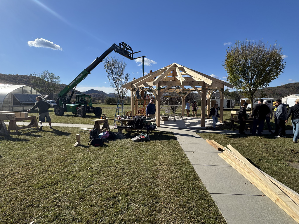 A panoramic shot of the timber framing project in progress. A large green telescopic forklift/telehandler is positioned to the left, assisting with lifting the heavy wooden beams for the gazebo roof structure. Students and adults are working around the frame, with mountains and a greenhouse visible in the background.
