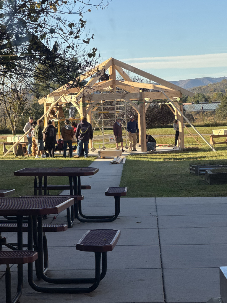 A wider view showing a large group of students and adults constructing a wooden timber frame pavilion. The structure is mostly complete, featuring a hipped roof frame. Picnic tables are in the foreground, and mountains are visible in the distant background.