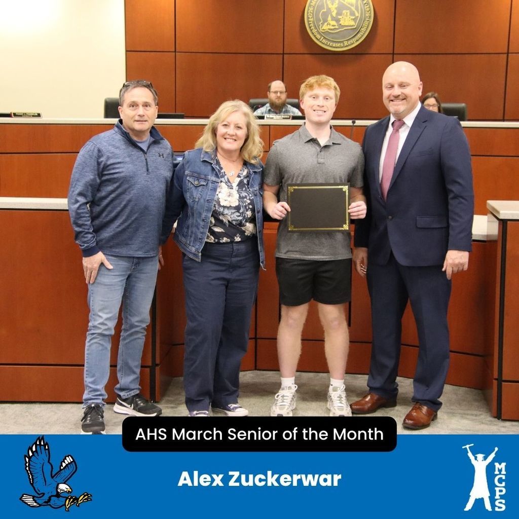 A high school student, Alex Zuckerwar, stands in a wood-paneled boardroom holding a black certificate. He is flanked by two adults on his left and a man in a suit on his right. The bottom banner identifies him as the AHS March Senior of the Month.