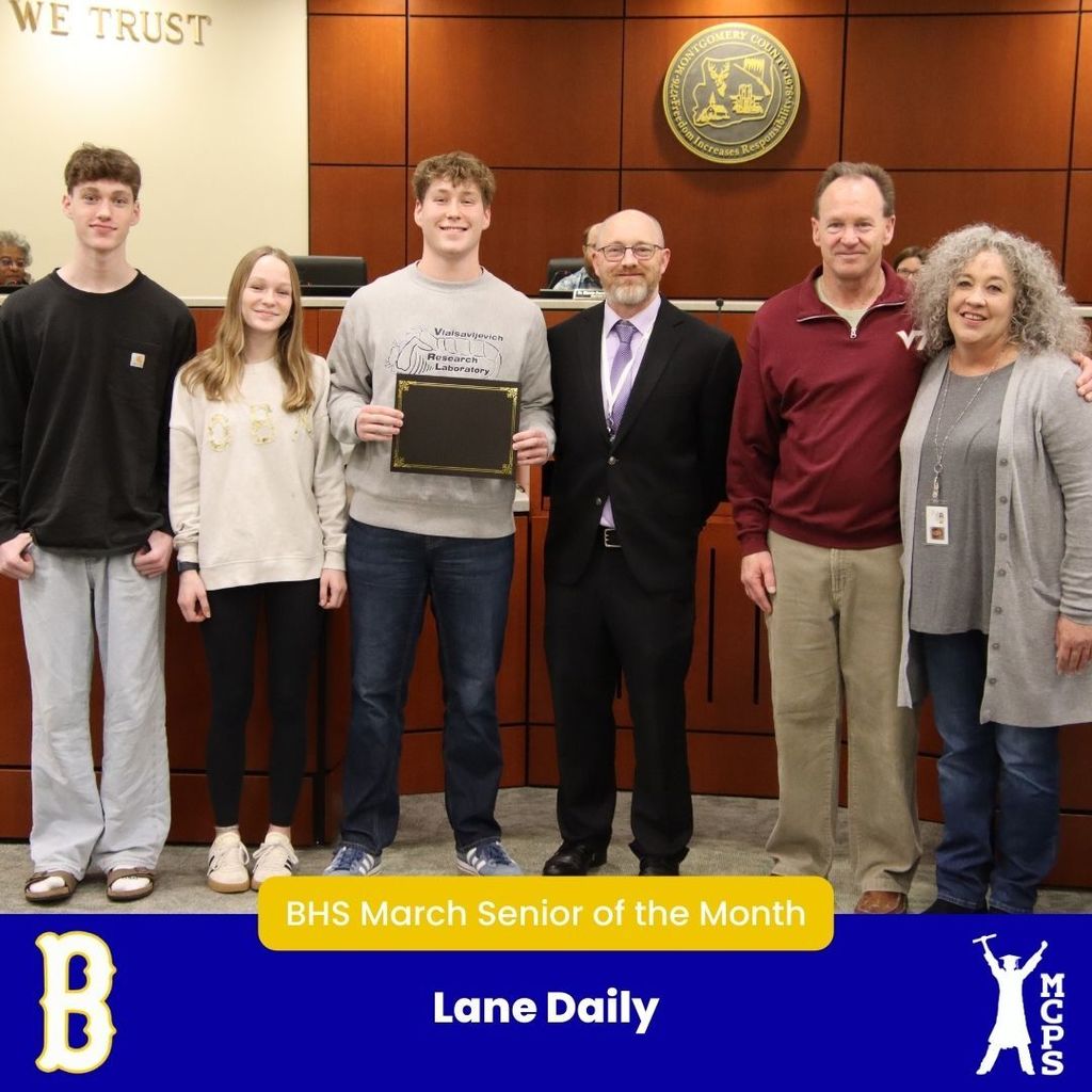 A group of six people stand in a boardroom. In the center, a student named Lane Daily holds a certificate. He is joined by two other teenagers and three adults. The bottom banner identifies him as the BHS March Senior of the Month.