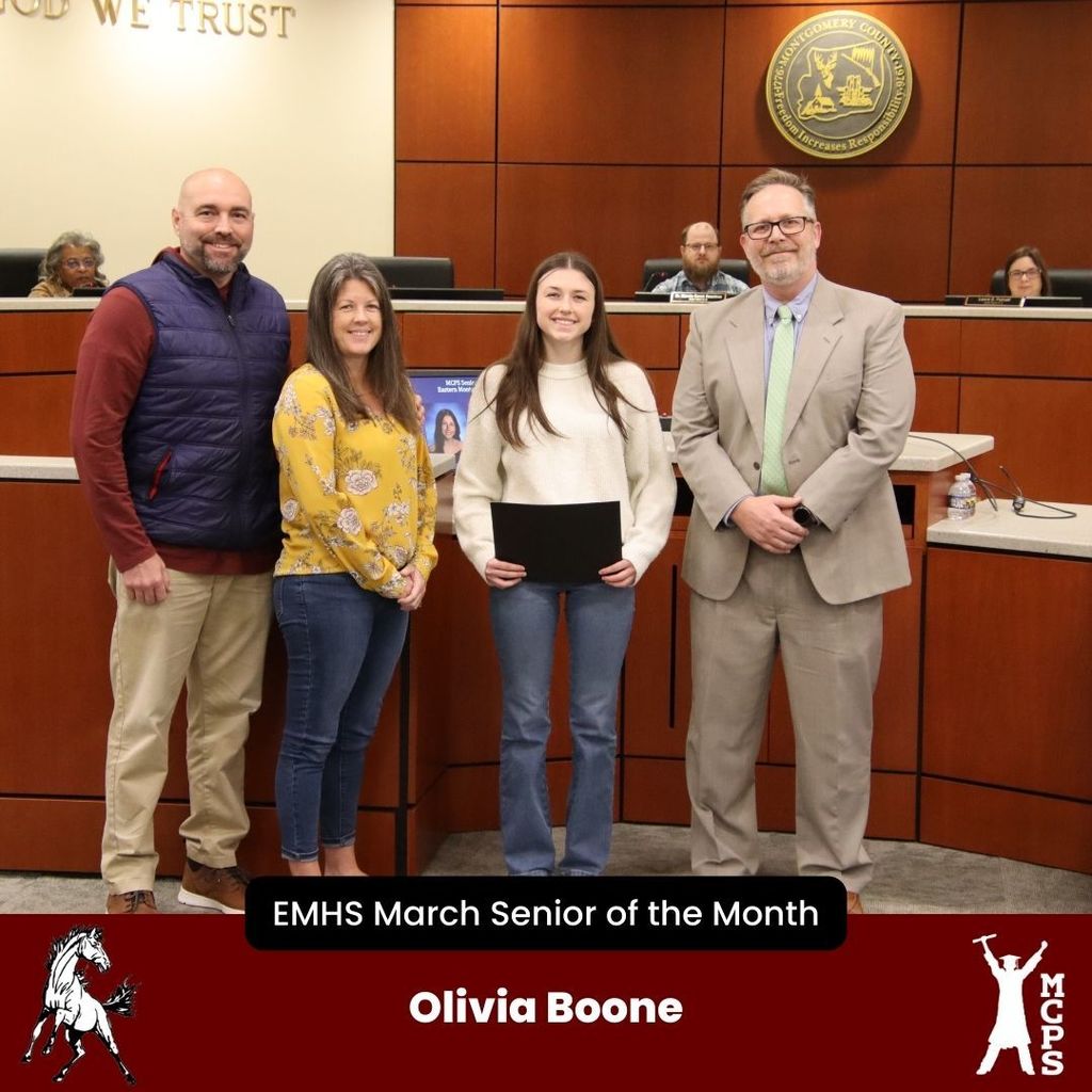 A student, Olivia Boone, stands in a boardroom holding a black certificate. She is accompanied by two adults on her left and a man in a tan suit and green tie on her right. The bottom banner identifies her as the EMHS March Senior of the Month.