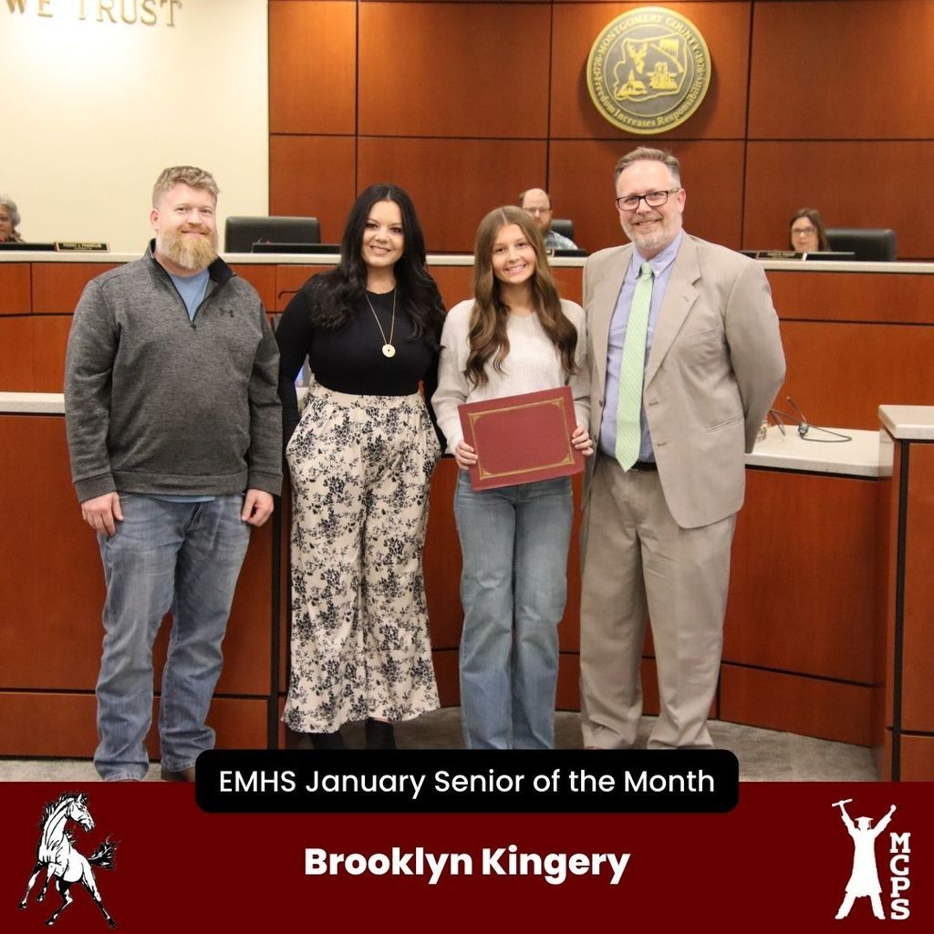 A student, Brooklyn Kingery, stands in a boardroom holding a red certificate. She is flanked by two adults on her left and a man in a tan suit on her right. The bottom banner identifies her as the EMHS January Senior of the Month.