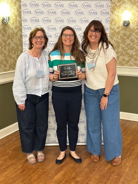 Three women stand smiling in front of a white "VaASL" step-and-repeat backdrop. The woman in the center, Jennifer Singleton, wears a striped blue, green, and white shirt and holds a black and gold plaque that reads "Roanoke Regional School Librarian of the Year." The woman on the left wears a light blue blouse and dark jeans, and the woman on the right wears a white eyelet top and wide-leg denim pants.