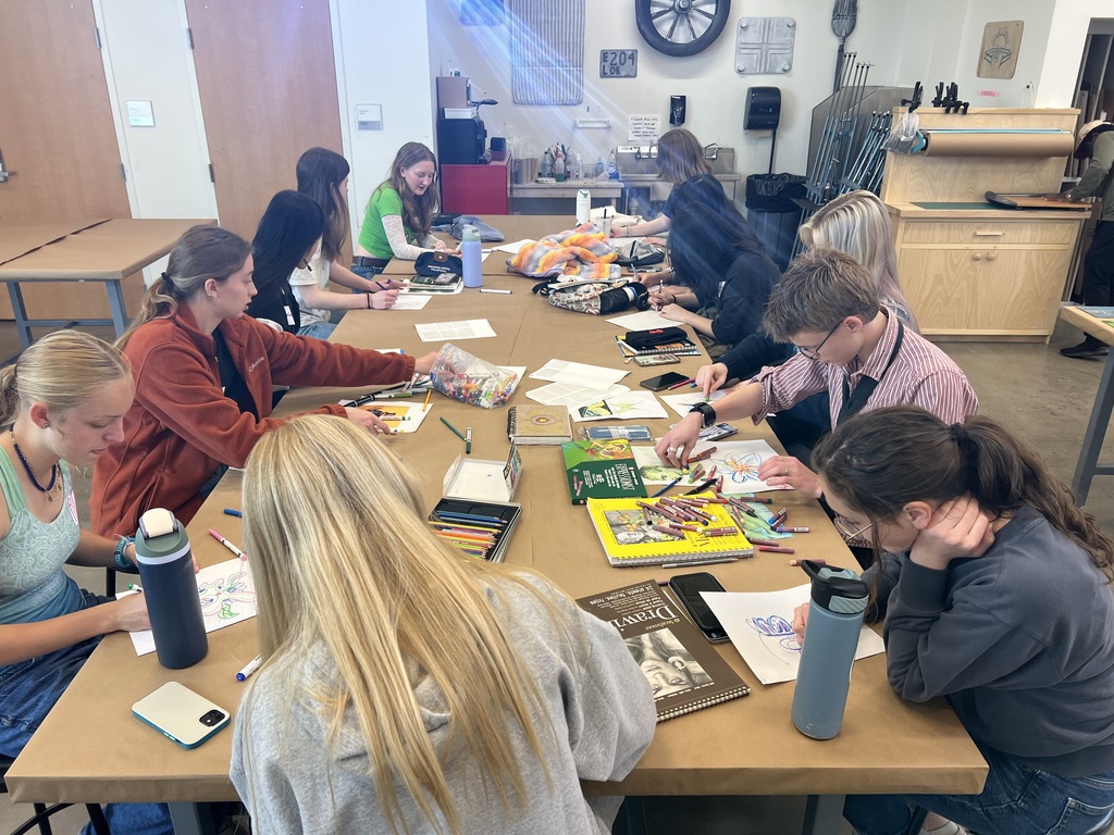 High school students huddled together and sitting at long tables, using markers and colored pencils to brainstorm and sketch initial designs on paper.