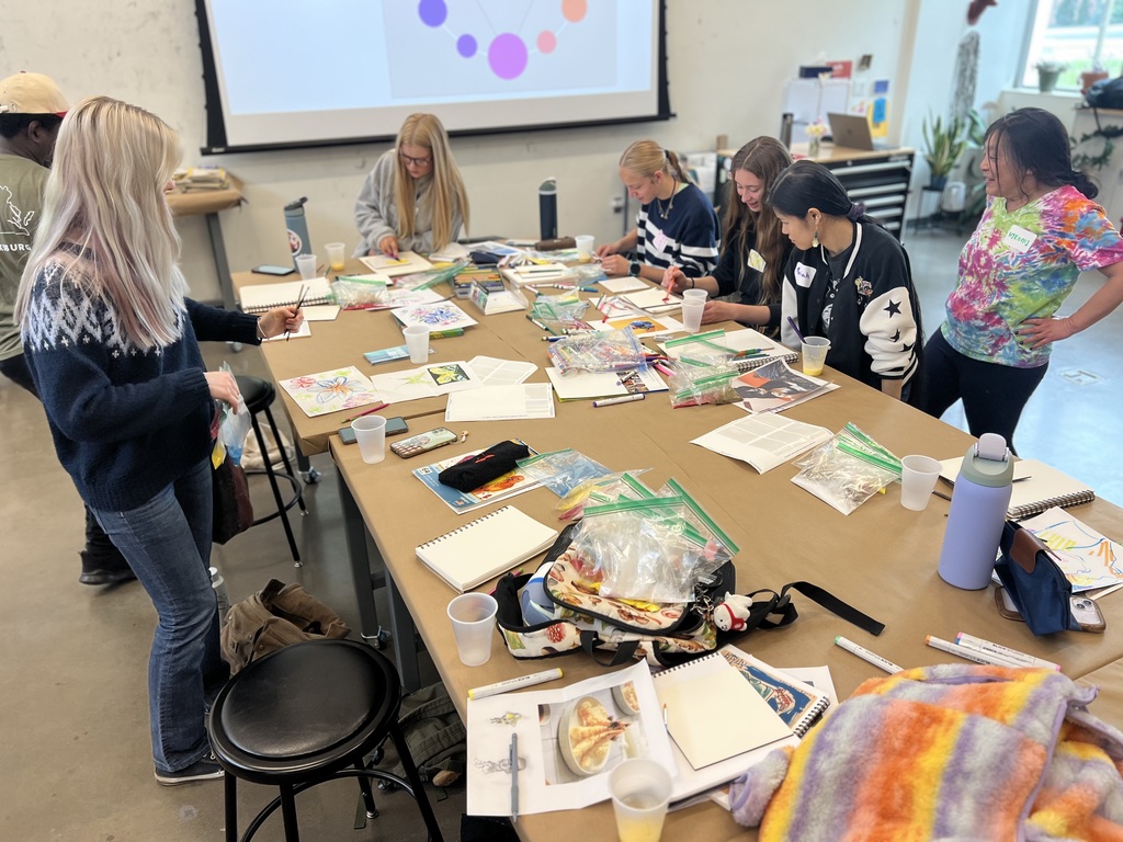 Students working with Professor Hiromi Okumura. They are using small plastic cups of yellow egg-yolk mixture and various pigments to paint butterflies and portraits on white panels. A color wheel is projected in the background.