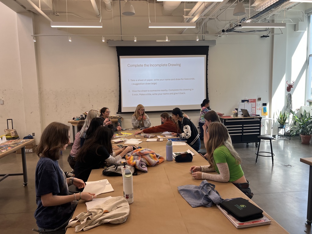 A wide shot of a bright, modern art studio at Virginia Tech. High school students sit at long tables covered in brown paper, looking at a projector screen that reads "Complete the Incomplete Drawing."