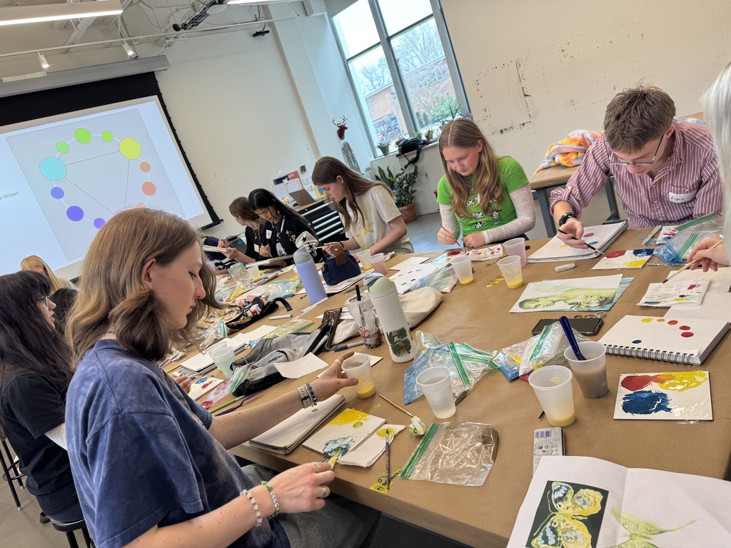 Close-up bird's-eye view of the workstation. You can see the handmade tempera paint in cups, vibrant color studies, and detailed paintings of landscapes, tigers, and floral patterns in progress.