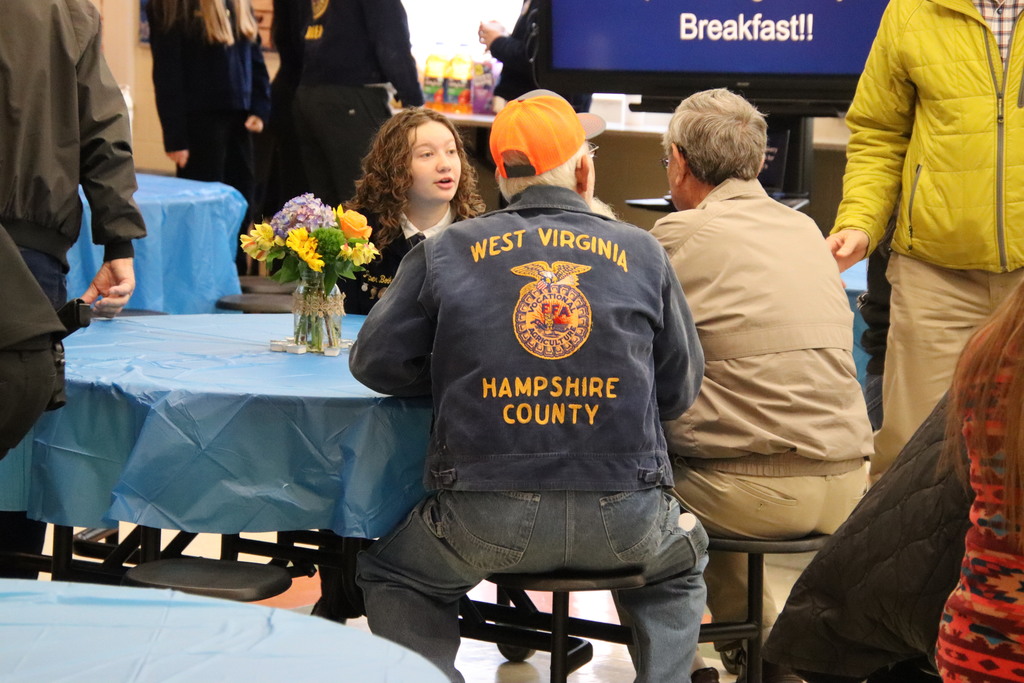 A Riner FFA student and an older farmer in a West Virginia FFA Alumni jacket share a smile and conversation over a blue-clothed table.