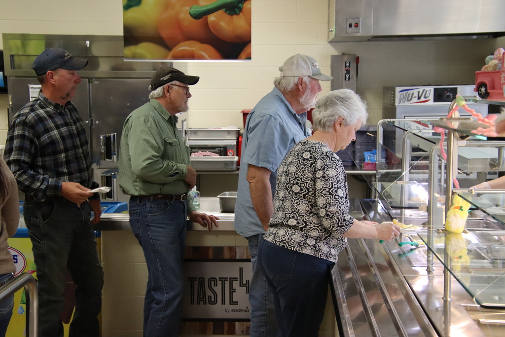 Local farmers in plaid shirts and caps move through the school cafeteria serving line to get their breakfast.