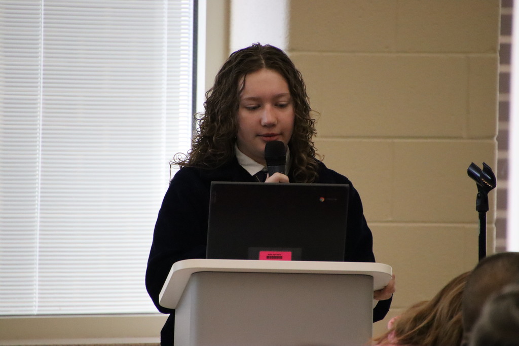 A Riner FFA member with curly hair speaks confidently into a microphone at a podium during the breakfast presentation.