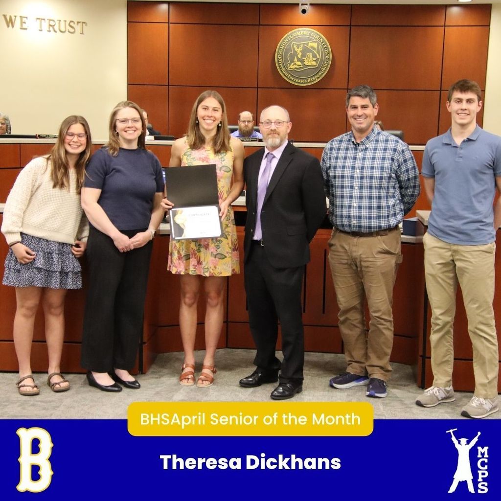 A formal group photo titled "BHS April Senior of the Month" featuring Theresa Dickhans. Theresa stands in the center holding a white certificate, wearing a yellow floral dress. She is flanked by four family members and a man in a black suit. The photo is taken in a boardroom with a circular seal on the wooden wall. A large yellow "B" mascot and the MCPS logo are at the bottom.