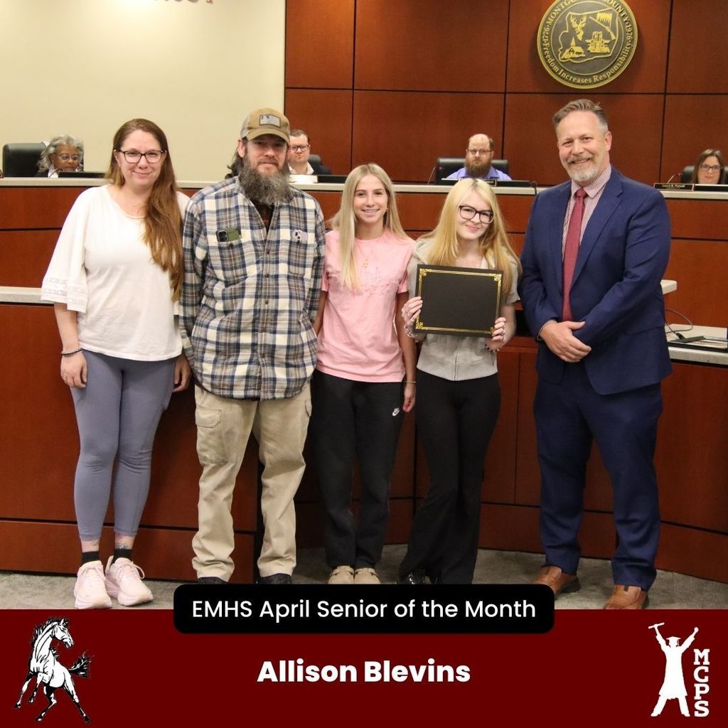 A formal group photo titled "EMHS April Senior of the Month" featuring Allison Blevins. Allison stands on the right-center holding a black award certificate. She is accompanied by three family members and a school official in a navy blue suit. The background shows a boardroom setting with a wooden wall and seal. A white mustang mascot and the MCPS logo are at the bottom.