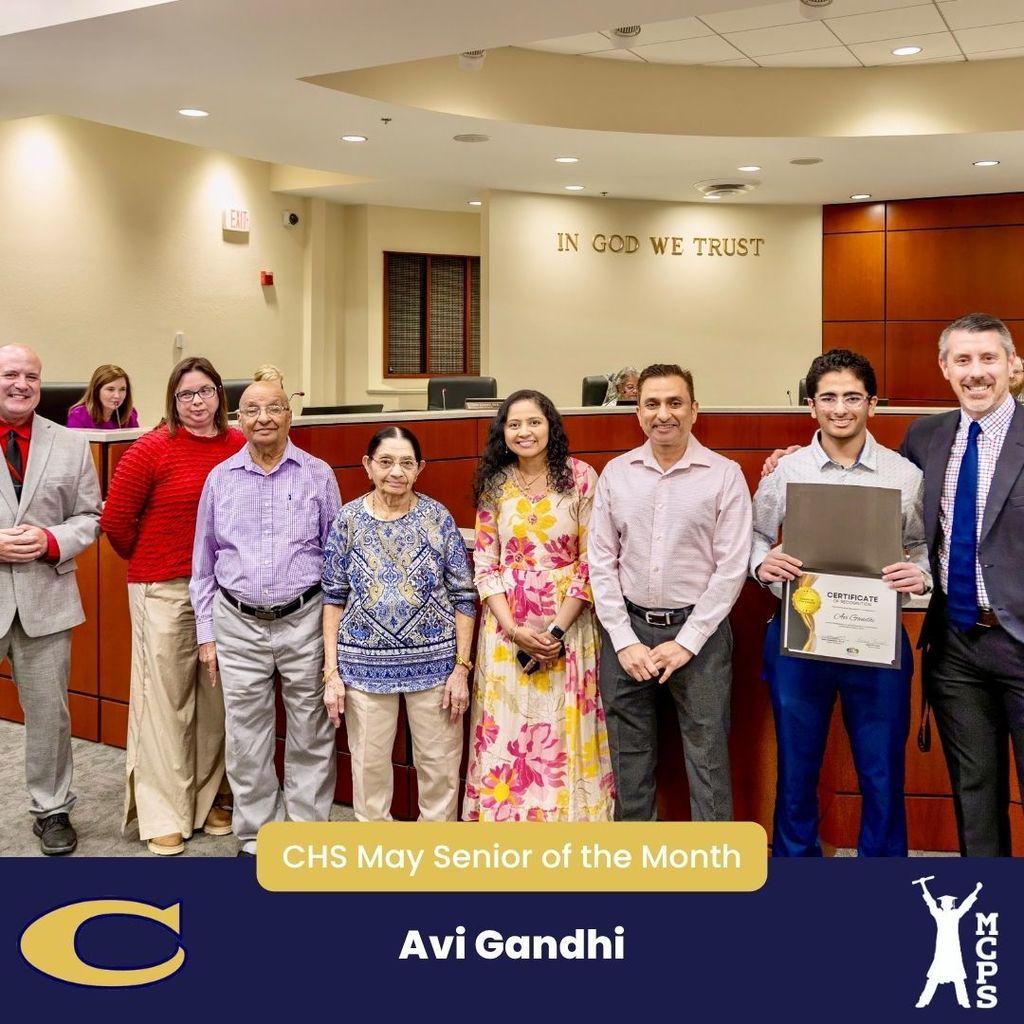 Avi Gandhi holds his CHS May Senior of the Month certificate, surrounded by family and school officials in a wood-paneled room