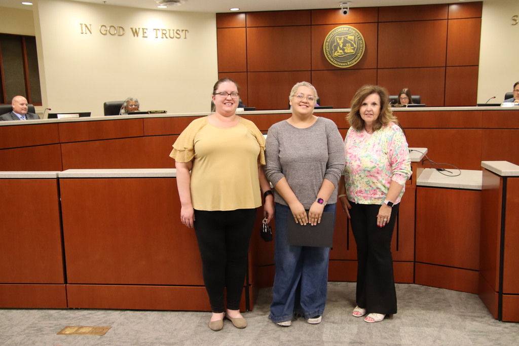 Three women stand smiling in a government chambers room with wood-paneled walls, a dais, and the words "IN GOD WE TRUST" on the wall behind them. The woman on the left wears a yellow top and black pants. The student in the center has short blonde hair, wears a grey long-sleeve shirt, wide-leg jeans, and holds a black certificate folder. The woman on the right wears a pink floral patterned cardigan and black pants.