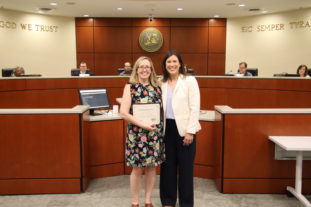 Two women stand smiling together in a government chambers room with wood-paneled walls, a curved dais, and committee members seated in the background. The phrases "GOD WE TRUST" and "SIC SEMPER TYRANNIS" are visible on the wall. The woman on the left has blonde hair, wears glasses, and a black sleeveless dress with a colorful floral pattern, and holds an award certificate. The woman on the right has dark brown hair and wears a white blazer over a light blue top with dark navy wide-leg trousers.