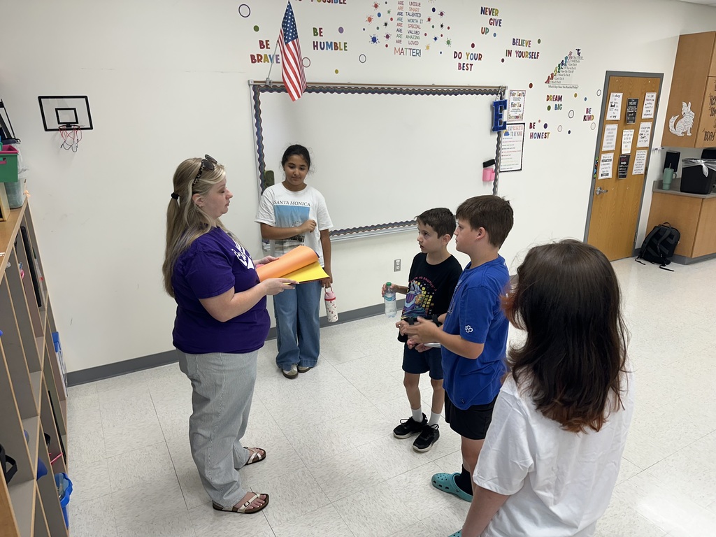 A classroom scene showing a female teacher in a purple shirt standing and holding a stack of colorful construction paper. She is speaking to a group of four young students standing in front of her. The classroom features a whiteboard decorated with motivational words like "Be Brave," "Be Humble," and "Do Your Best," along with a small American flag mounted on top.