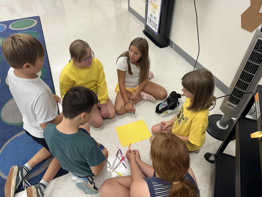 A high-angle shot of six young students sitting tightly in a circle on the floor of a classroom. In the middle of the group is a bright yellow sheet of paper surrounded by several markers scattered on the tiles. A few of the children are wearing yellow shirts, and they appear to be collaborating or discussing an art project together.