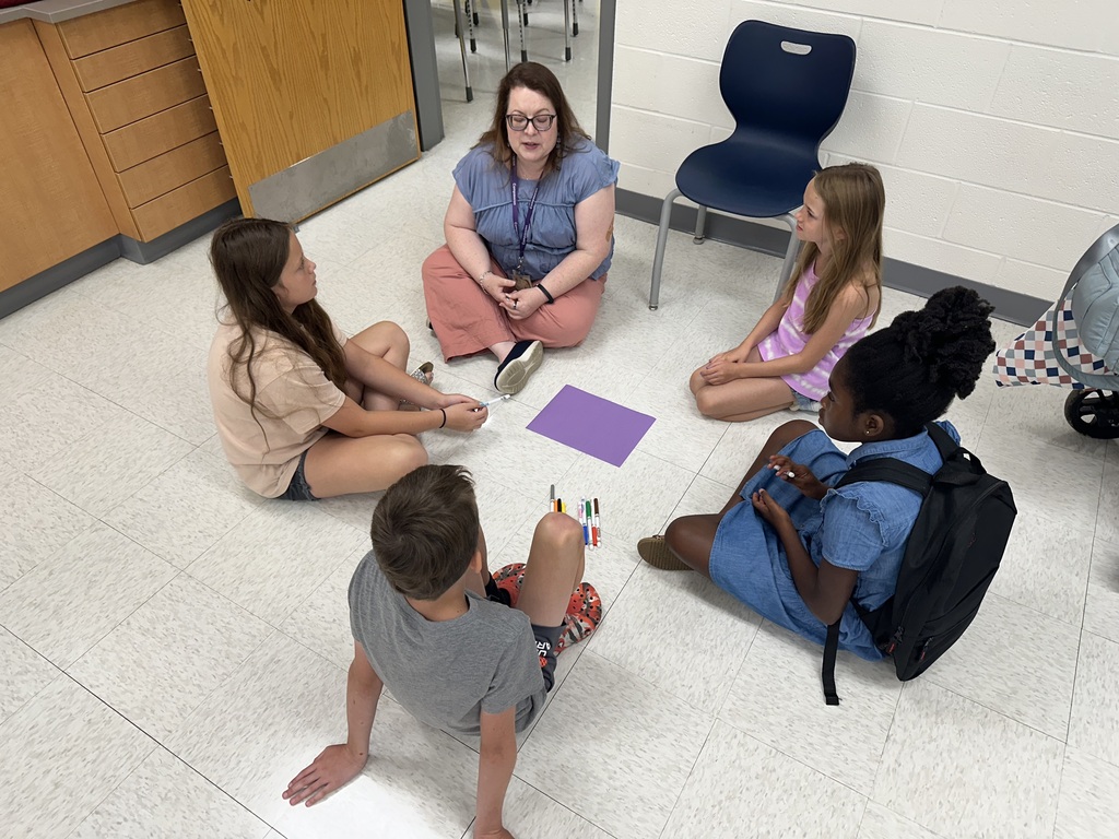 An overhead shot of an adult woman and four young students sitting cross-legged in a circle on a tiled classroom floor. The woman wears glasses, a blue shirt, and pink pants. The children consist of three girls and one boy. In the center of the circle lies a purple sheet of paper and a small pile of colored markers. One girl holds a marker while looking toward the woman, who appears to be speaking or leading an activity.