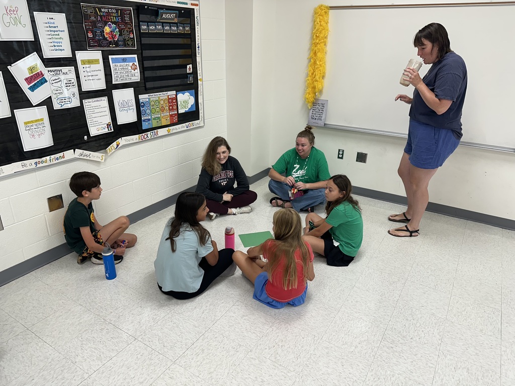 A group of five students and two adult women are gathered in a classroom corner near a large bulletin board. Four students and two adults sit cross-legged in a circle on the floor around a green sheet of paper. Another adult stands to the right, leaning against a white whiteboard while drinking from a large cup. The classroom walls are decorated with educational posters and a colorful calendar.