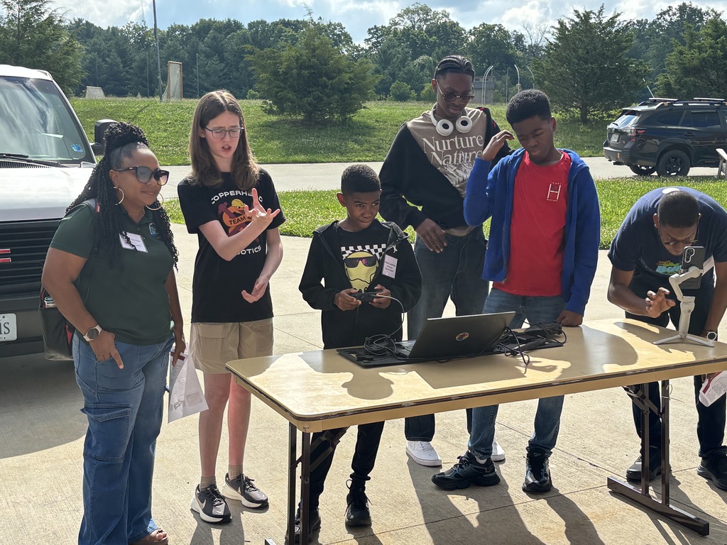 Students and mentors outdoors around a table as a young student uses a laptop and controller to operate a robotics demonstration.