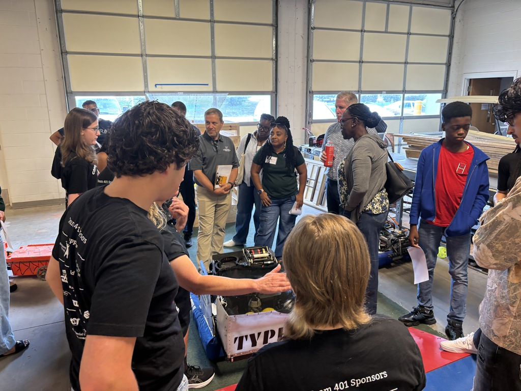 A diverse group of students and mentors gather in a workshop around a custom competition robot during a demonstration.