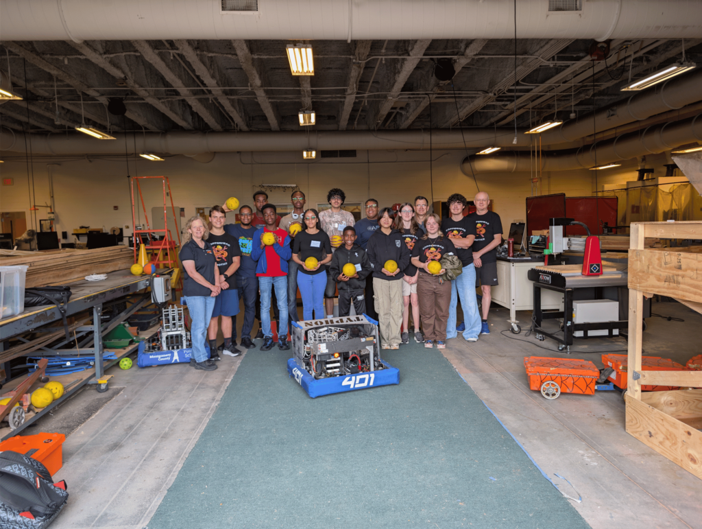A wide-angle, full group photograph taken from a distance, showing a diverse gathering of approximately 18 students and mentors standing on a long teal runner carpet that extends from the foreground back into a large, open-plan school robotics or shop classroom. They are lined up behind a large, custom-built industrial competition robot with "401" on its blue bumper. Several members hold bright yellow game balls. The room features high ceilings, exposed ventilation pipes, bright overhead fluorescent lighting, and is filled with other large machines, workbenches stacked with tools and parts, an orange crate, and other smaller robotics equipment, including another smaller robot on the left.