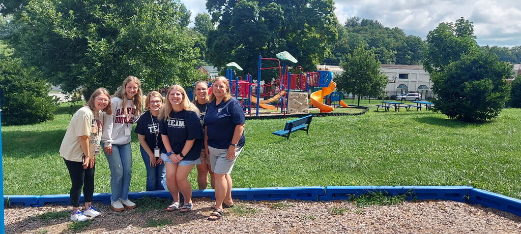 Six CES staff members stand together on the school playground with colorful play equipment, trees, and picnic tables in the background.
