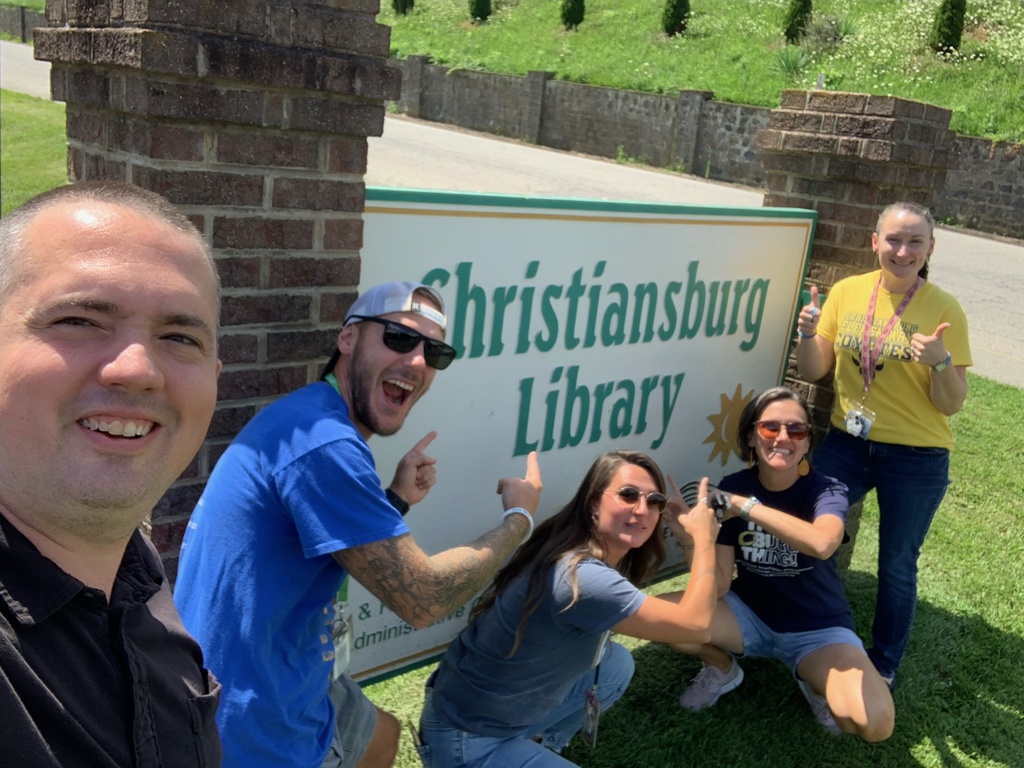 Five smiling CES staff members pose and point toward the Christiansburg Library sign on a bright, sunny day.