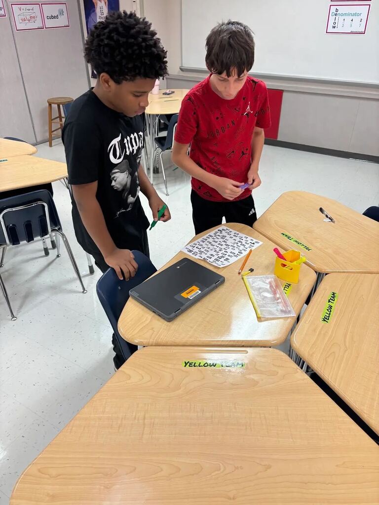 Two students work together at classroom desks with a number sheet, laptop, pencils, and colorful markers.