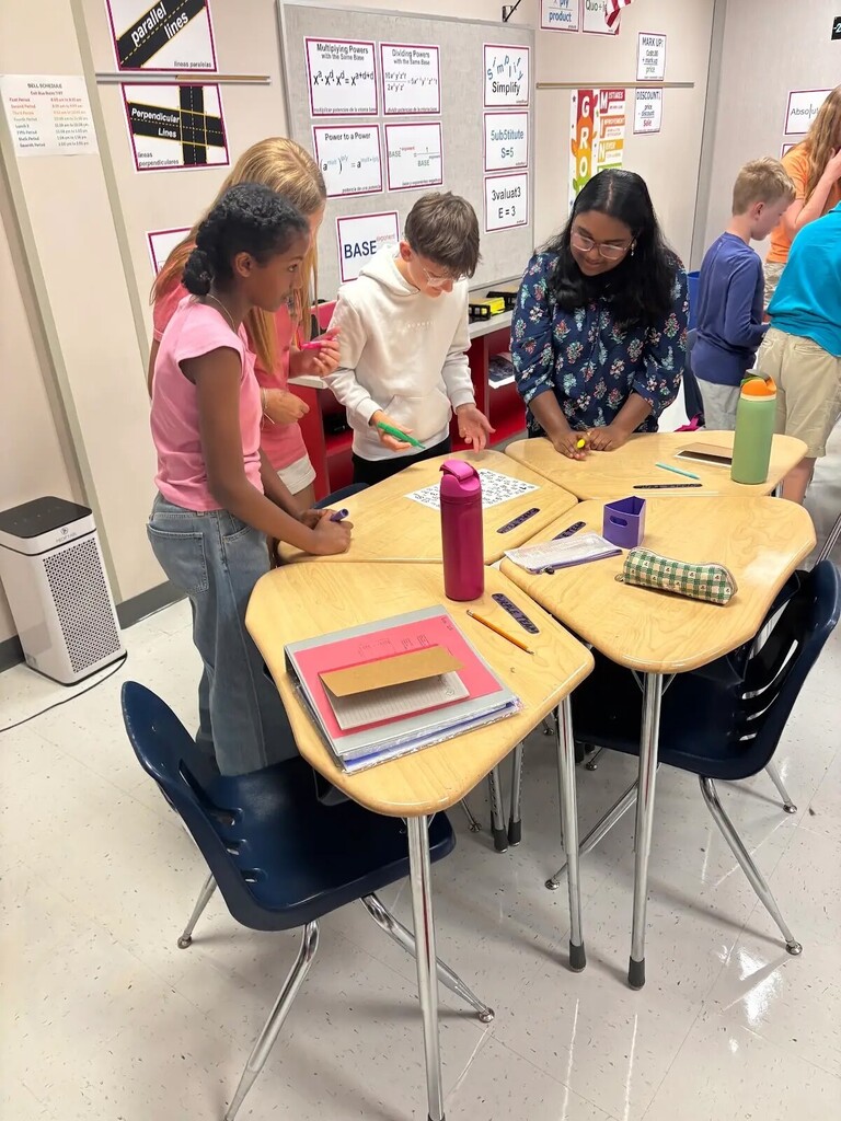 Students collaborate around classroom desks on a math activity using number sheets, markers, and school supplies.
