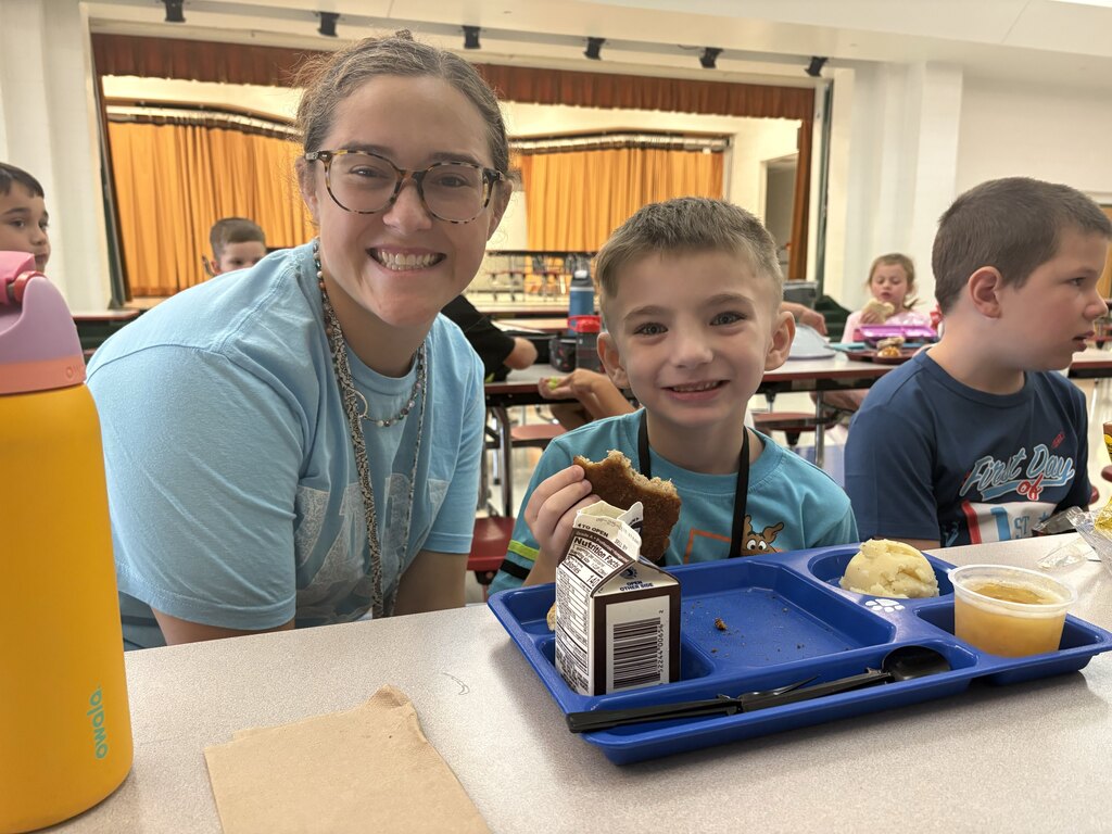 A smiling staff member sits beside a student eating lunch from a blue cafeteria tray on the first day of school.