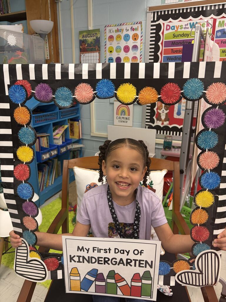A smiling kindergarten student poses inside a colorful classroom frame holding a “My First Day of Kindergarten” sign.