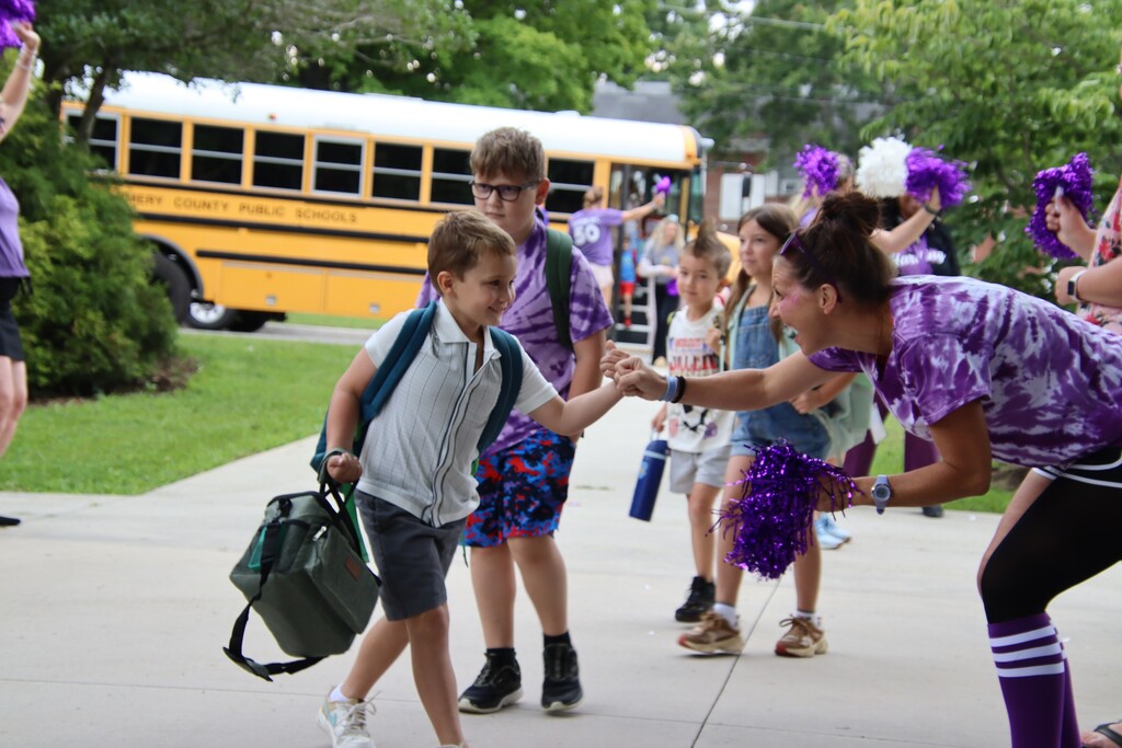 A student fist-bumps a cheering staff member holding purple pom-poms as students arrive from a school bus.