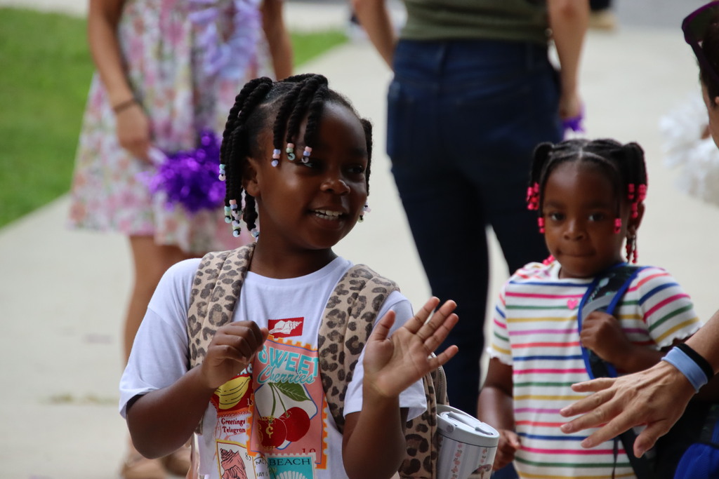 A smiling student with a backpack waves while arriving at school, with another student walking behind her.