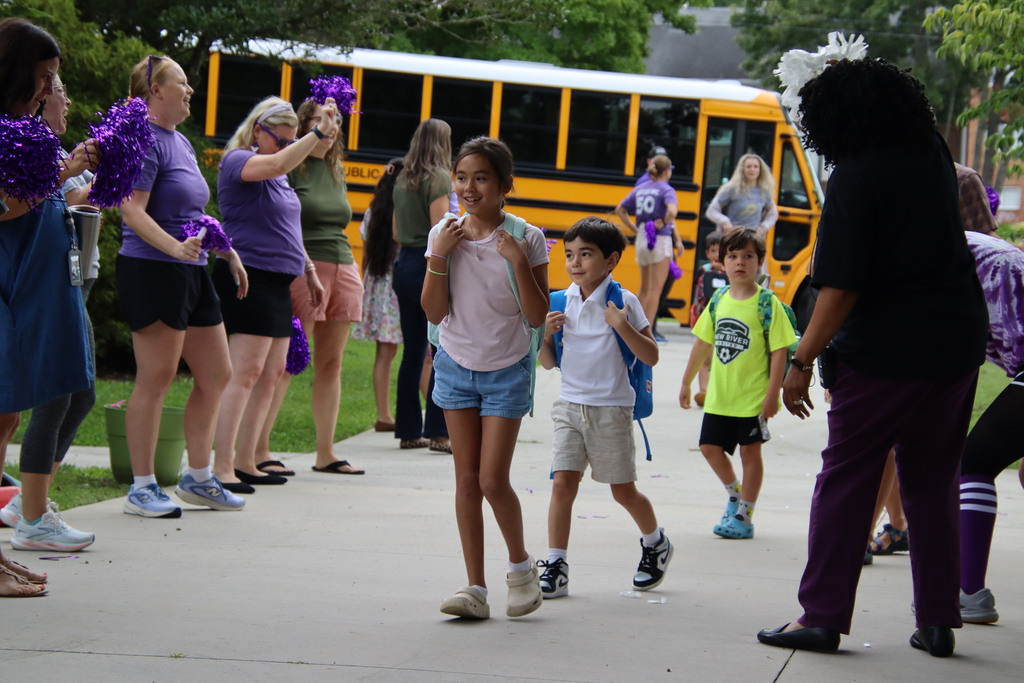 Students with backpacks walk from a yellow school bus as cheering staff welcome them with purple pom-poms.