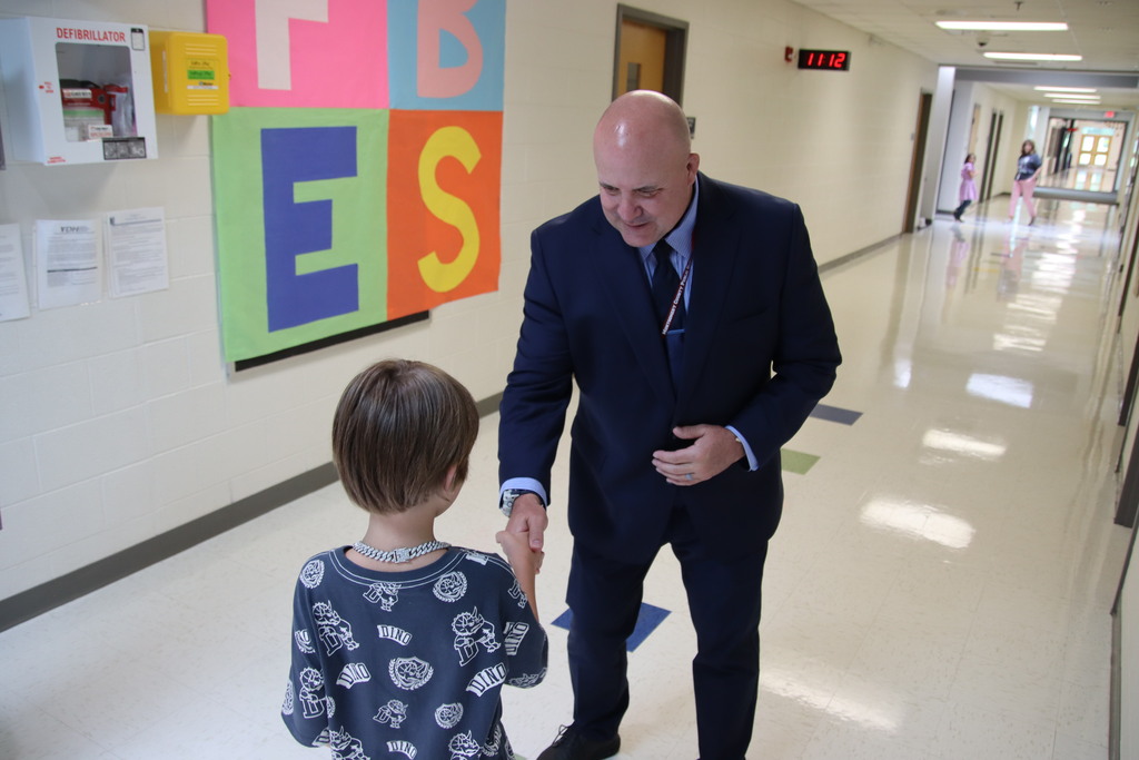 A school administrator in a suit shakes hands with a student in a bright elementary school hallway.