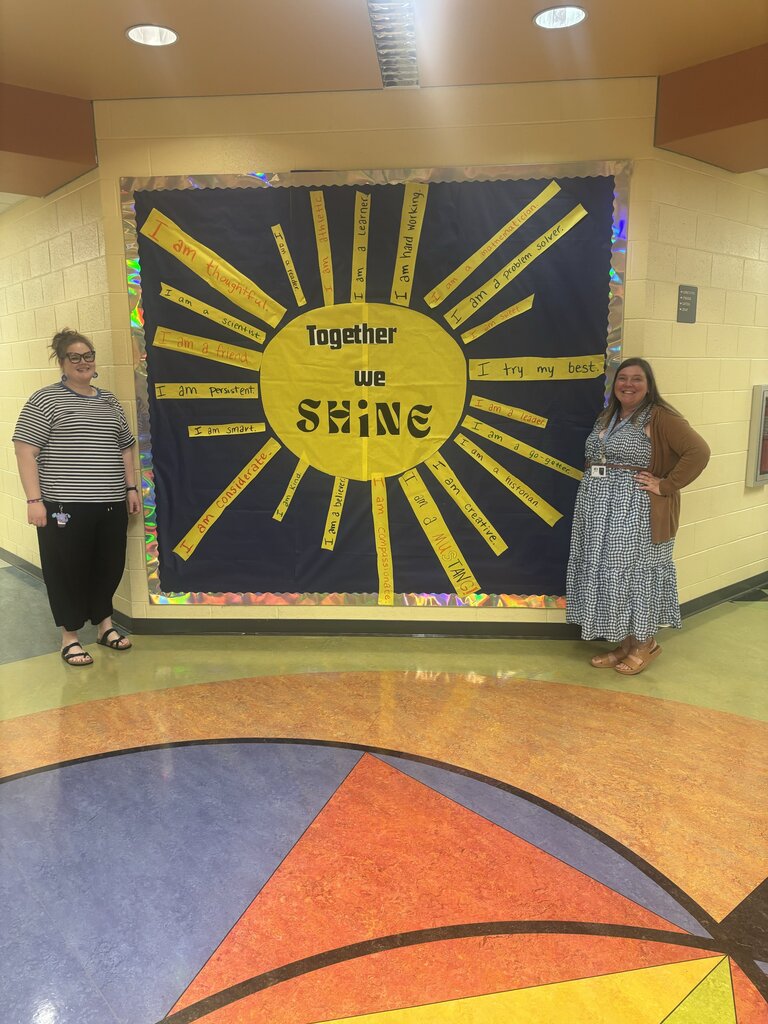 Two staff members stand beside a bulletin board featuring a large sun and the message, “Together we shine.”