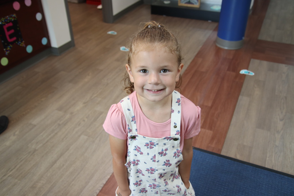 A smiling young student in floral overalls poses inside a school building on the first day of school.