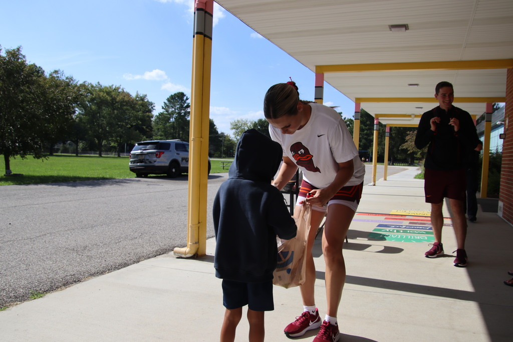 A student-athlete hands a bag of supplies to a child outside the school on a sunny day.