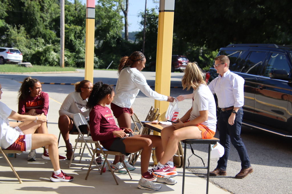 Virginia Tech student-athletes greet visitors and distribute supplies during a sunny outdoor school event.