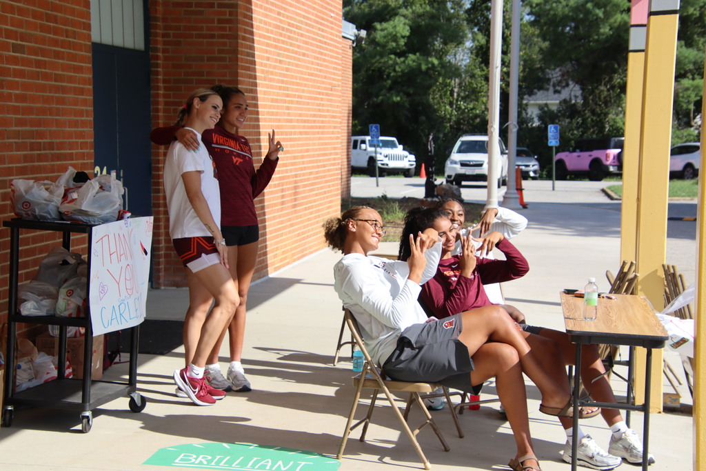 Student-athletes pose and relax near tables of donated supplies outside a brick school building.